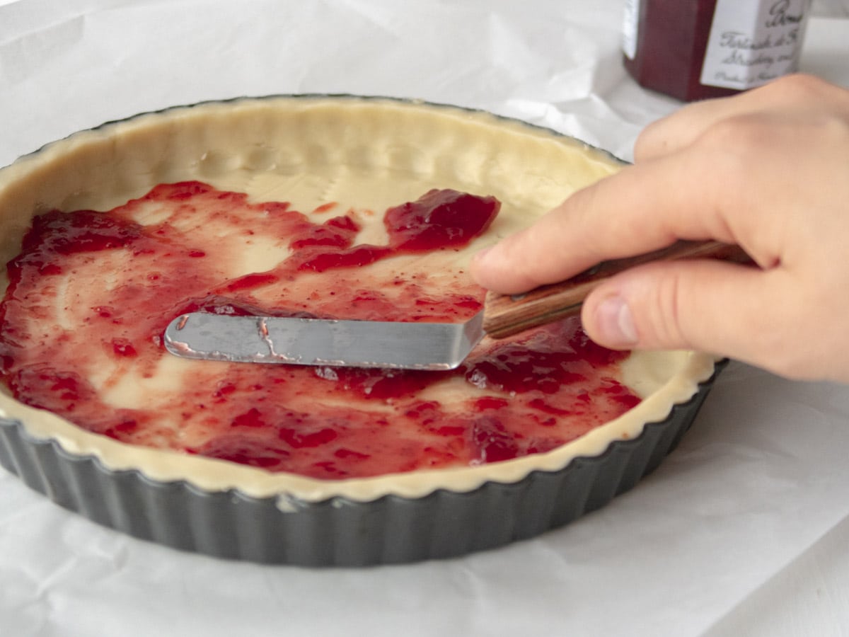 raw tart shell with strawberry jam being spread on the bottom