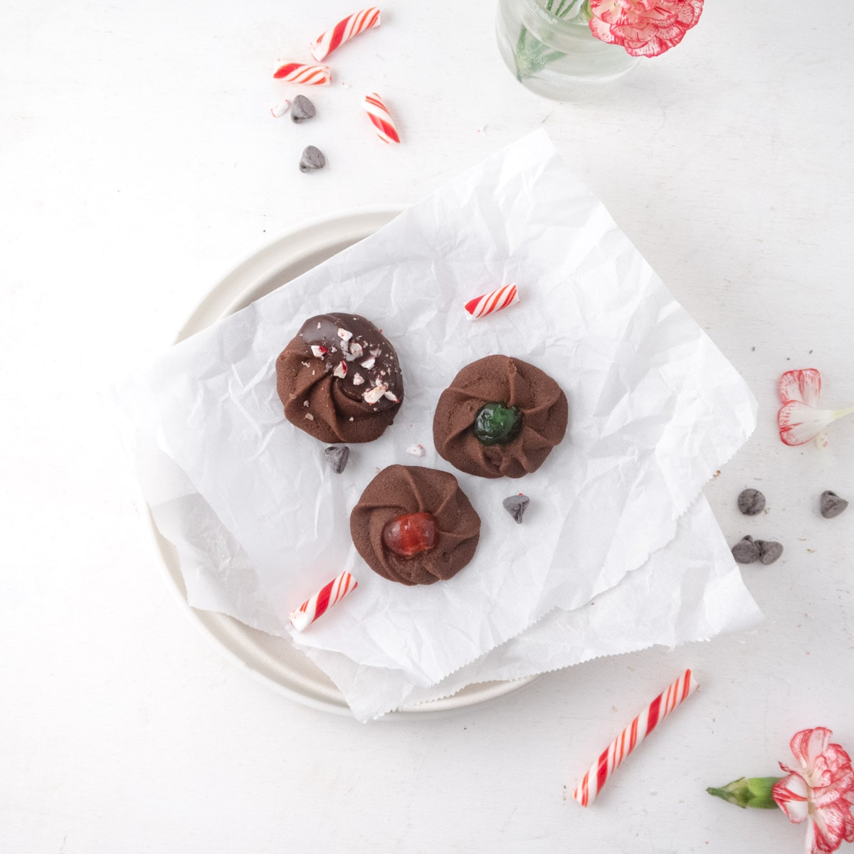 top view of 3 differently decorated chocolate butter cookies on a plate with some candy canes and flowers on the table