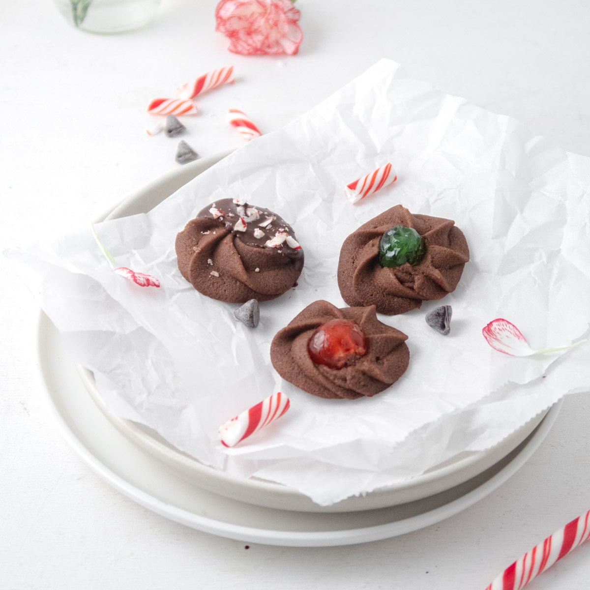 side view of 3 differently decorated chocolate butter cookies on a plate with some candy canes and flowers on the table