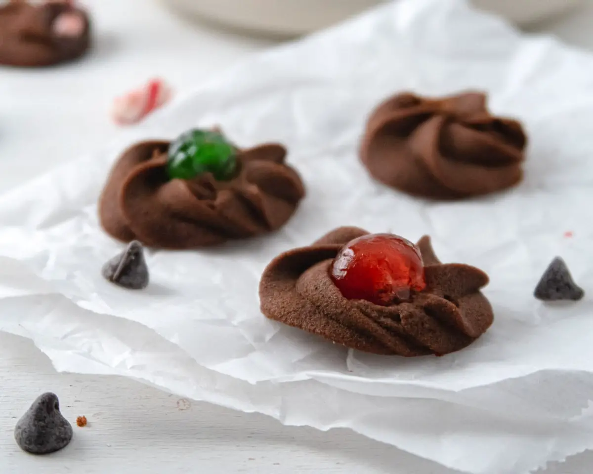 vegan chocolate butter cookies on top of a piece of crinkled parchment paper with a green and red cherry in the middle