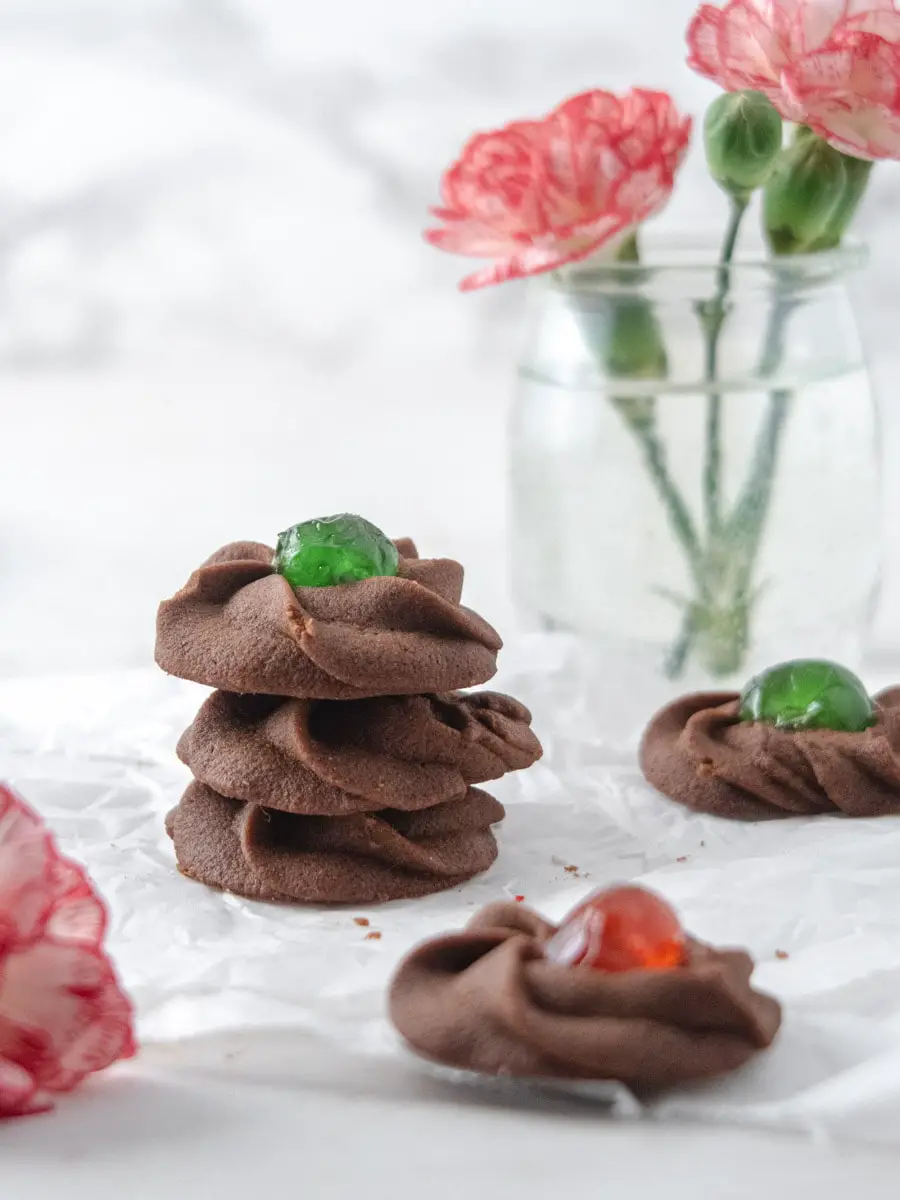 Chocolate cookies with a cheryr in the mibble stacked on top of each other with some flowers in the background