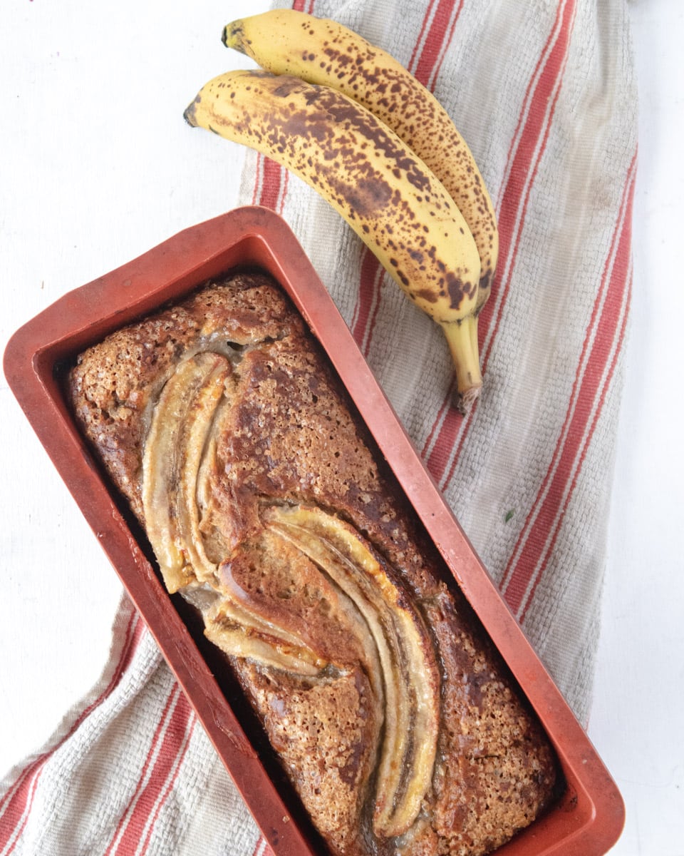 baked banana bread in a silicone pan on top of a red stripped dishtowel and bananas in the background