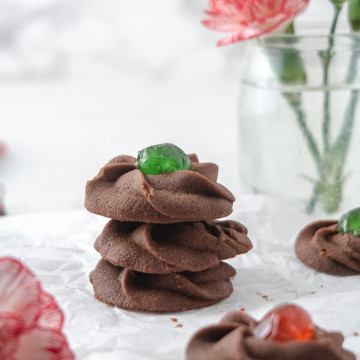 Chocolate cookies with a cheryr in the mibble stacked on top of each other with some flowers in the background