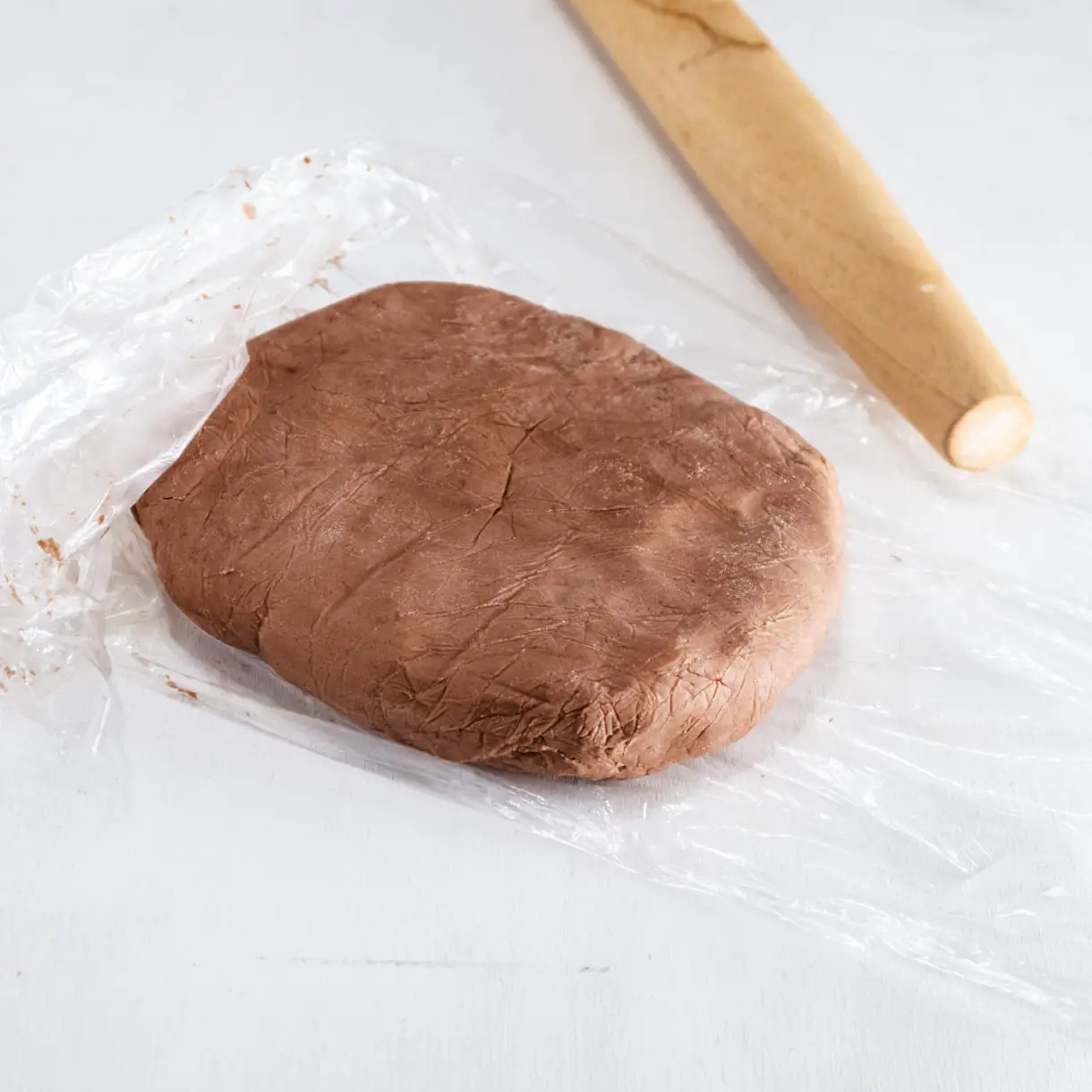 disk of cacao shortcrust dough on a piece of plastic wrap with a rolling pin next to it