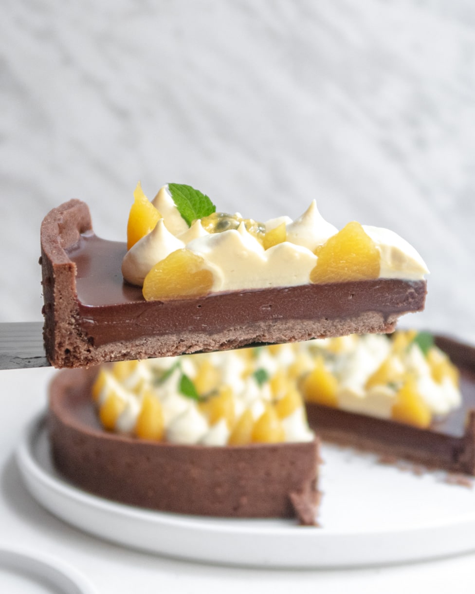 A slice of passionfruit chocolate tart held up in midair by a knife with the rest of the tart in the background