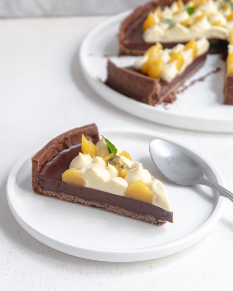 a slice of passionfruit and chocolate tart on a white plate with the whole tart in the background