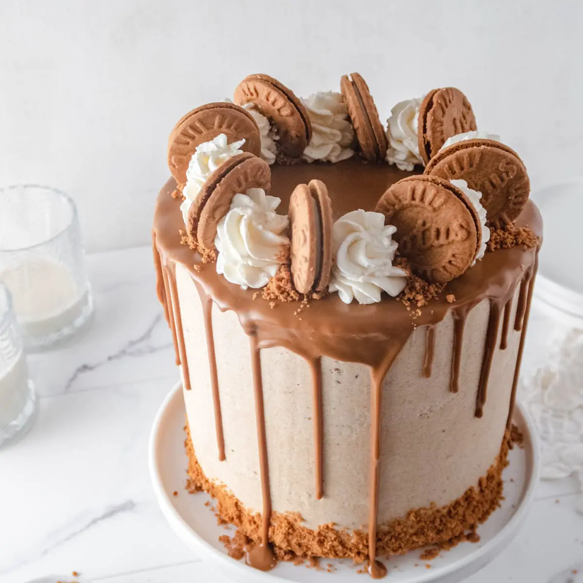 Angled down view of a vegan cake with a Biscoff drip and piped buttercream rosette and cookies on a white marbled background.