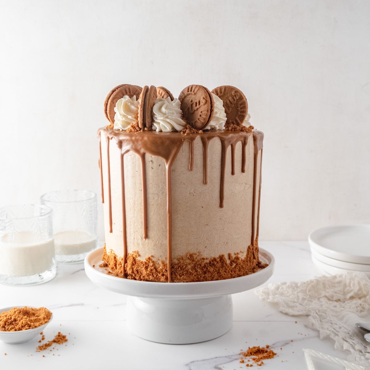 Vegan cake with a Biscoff drip and piped buttercream rosette and cookies on a cake stand in front of a white background with plates, glasses and cookies crumbles around.