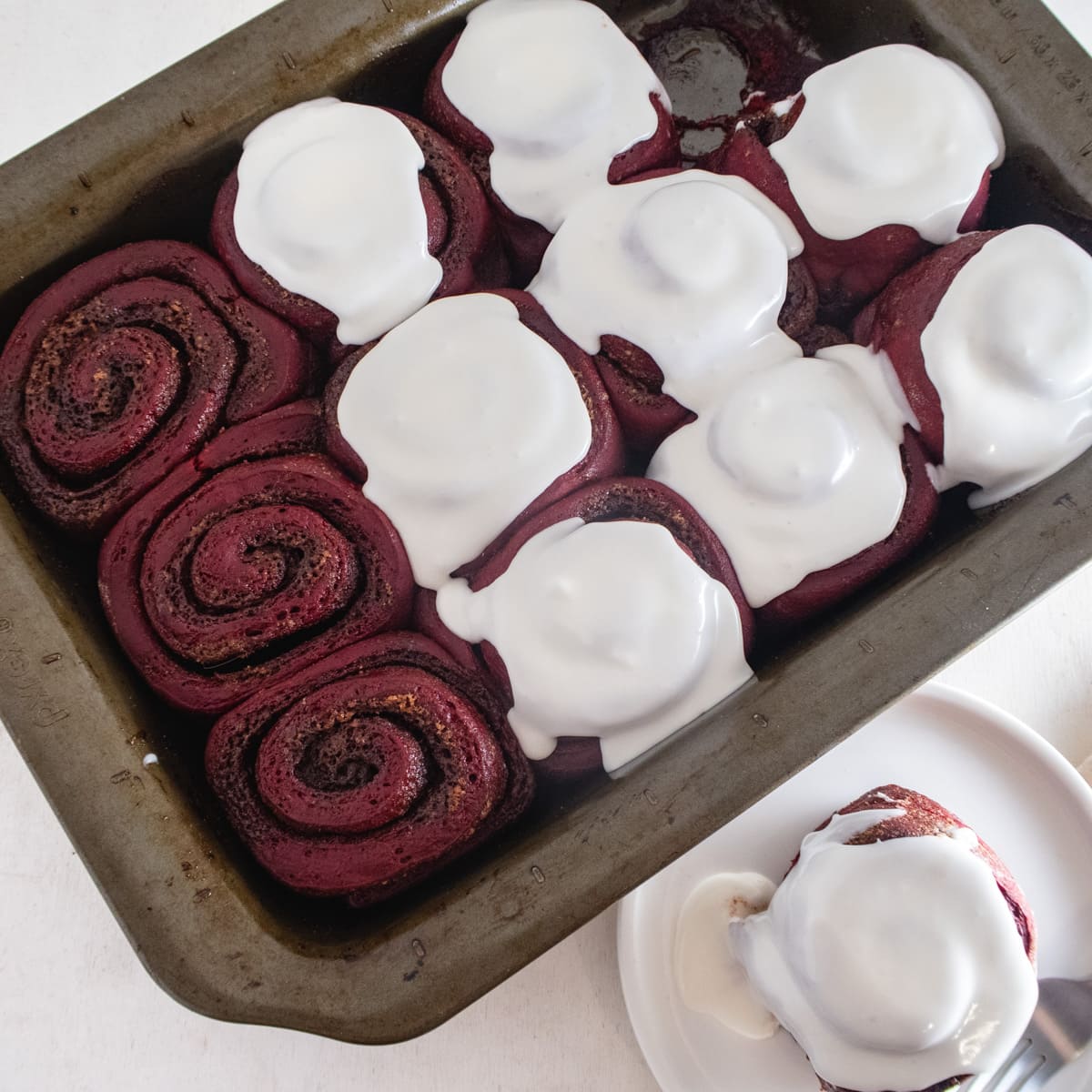 Overhead view of half iced vegan red velvet cinnamon rolls in a baking dish with a cinnamon an a plate next to it.