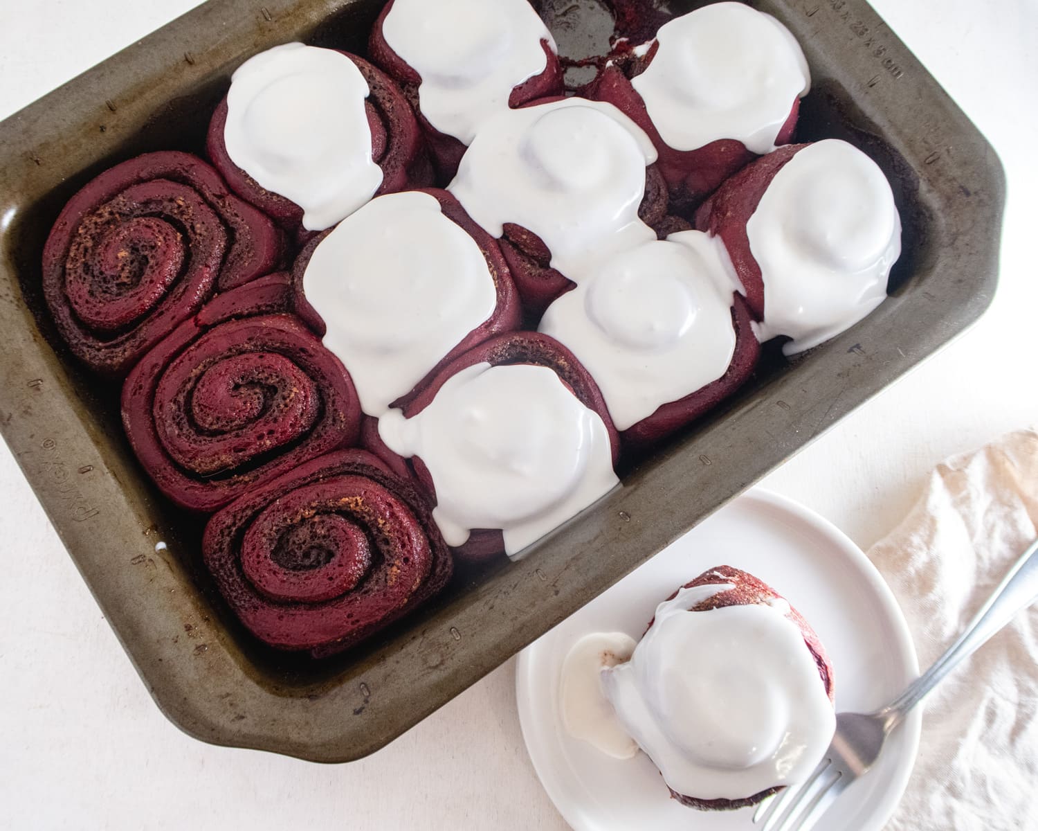 Overhead view of half iced vegan red velvet cinnamon rolls in a baking dish with a cinnamon an a plate next to it.