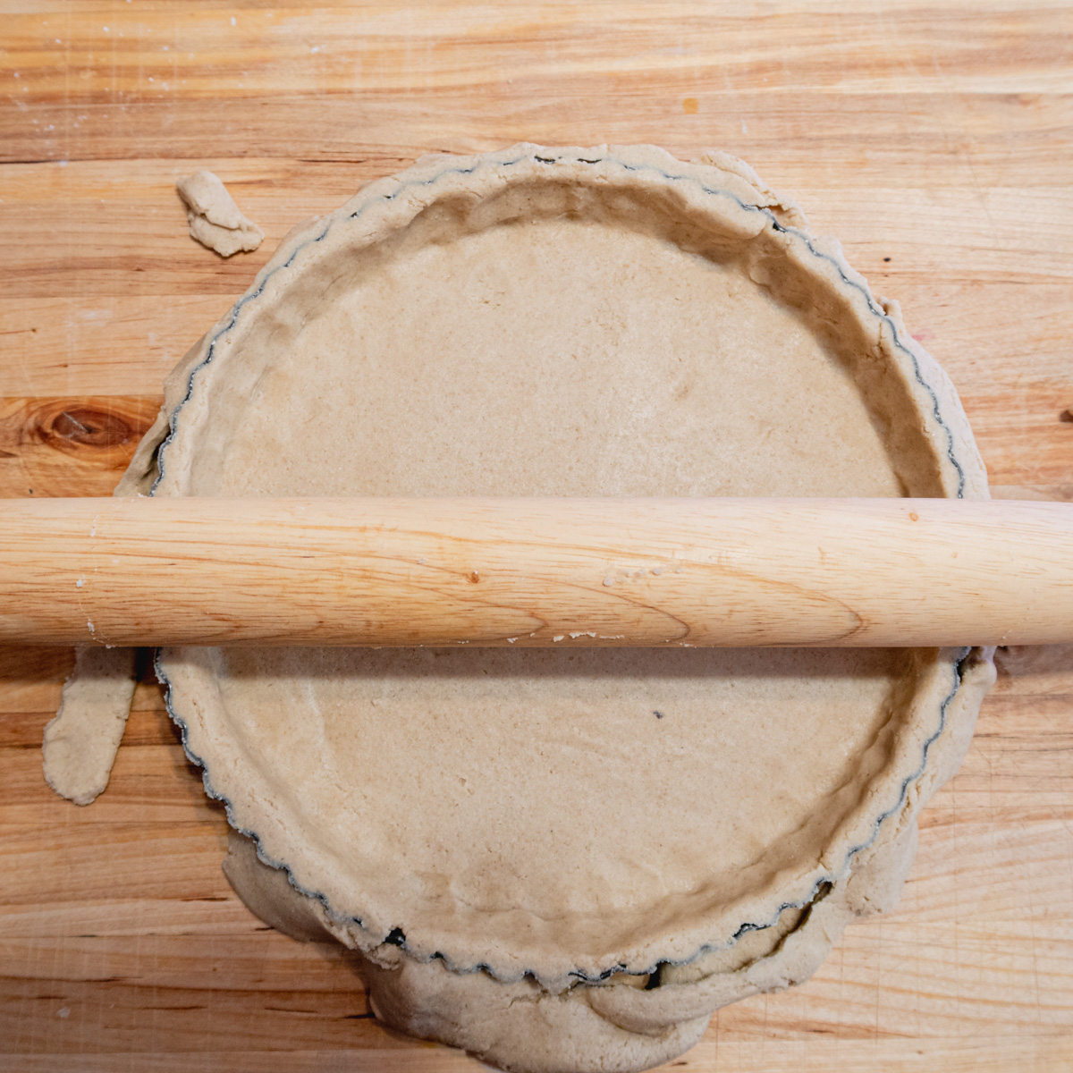 A rolling pin being rolled on the top edge of a dough-lined tart tin is cutting the excess dough.