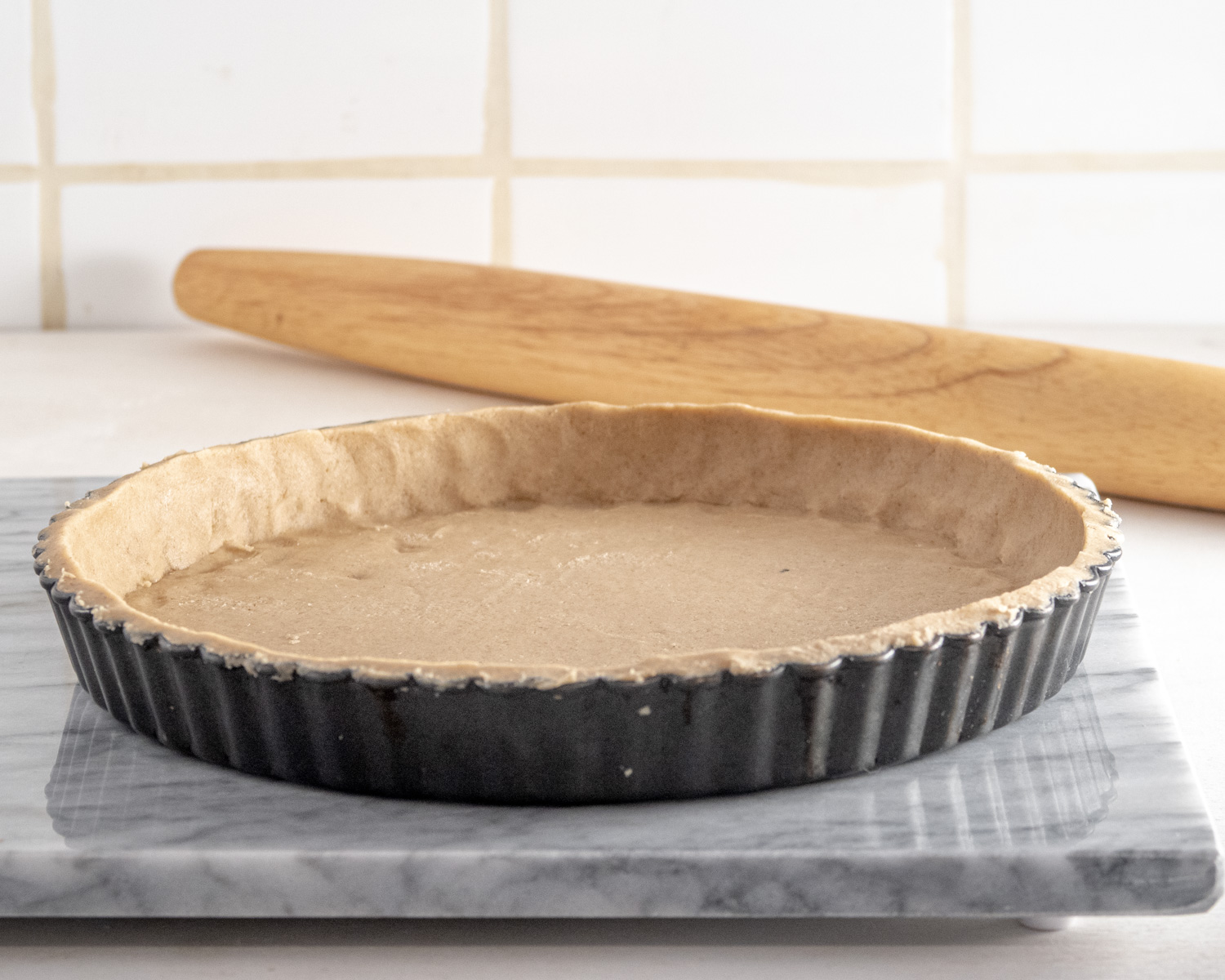 Side view of a raw gluten-free tart shell on a marble slab with a rolling pin in the background.