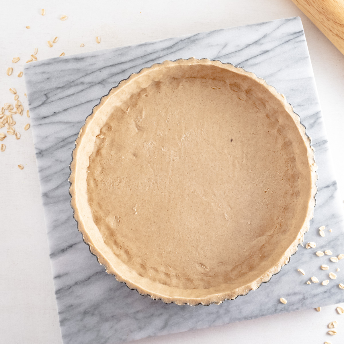 Raw gluten-free tart shell in a fluted tart tin on a slab of marble and oats around it.
