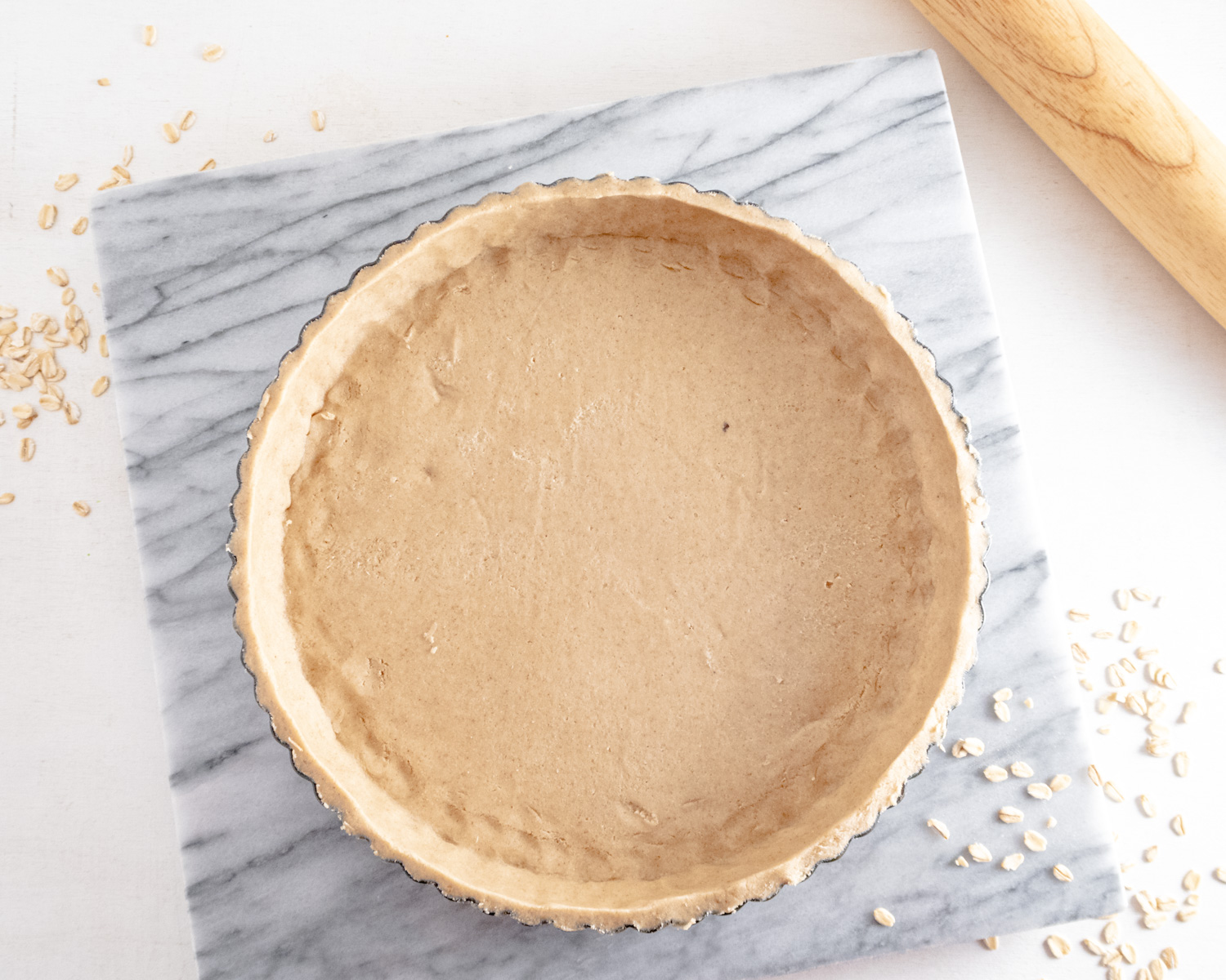 Raw gluten-free tart shell in a fluted tart tin on a slab of marble and oats around it.