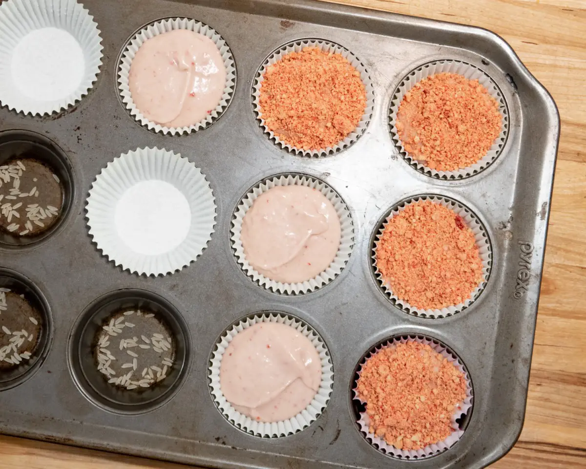 Cupcake pan in the process of being filled with strawberry cupcake batter and topped with strawberry crunch topping.