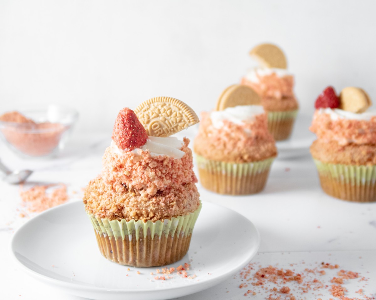 Single cupcake on a white plate with strawberry crunch topping and more cupcakes in the background. 