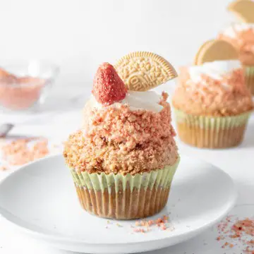 Single cupcake on a white plate with strawberry crunch topping and more cupcakes in the background.