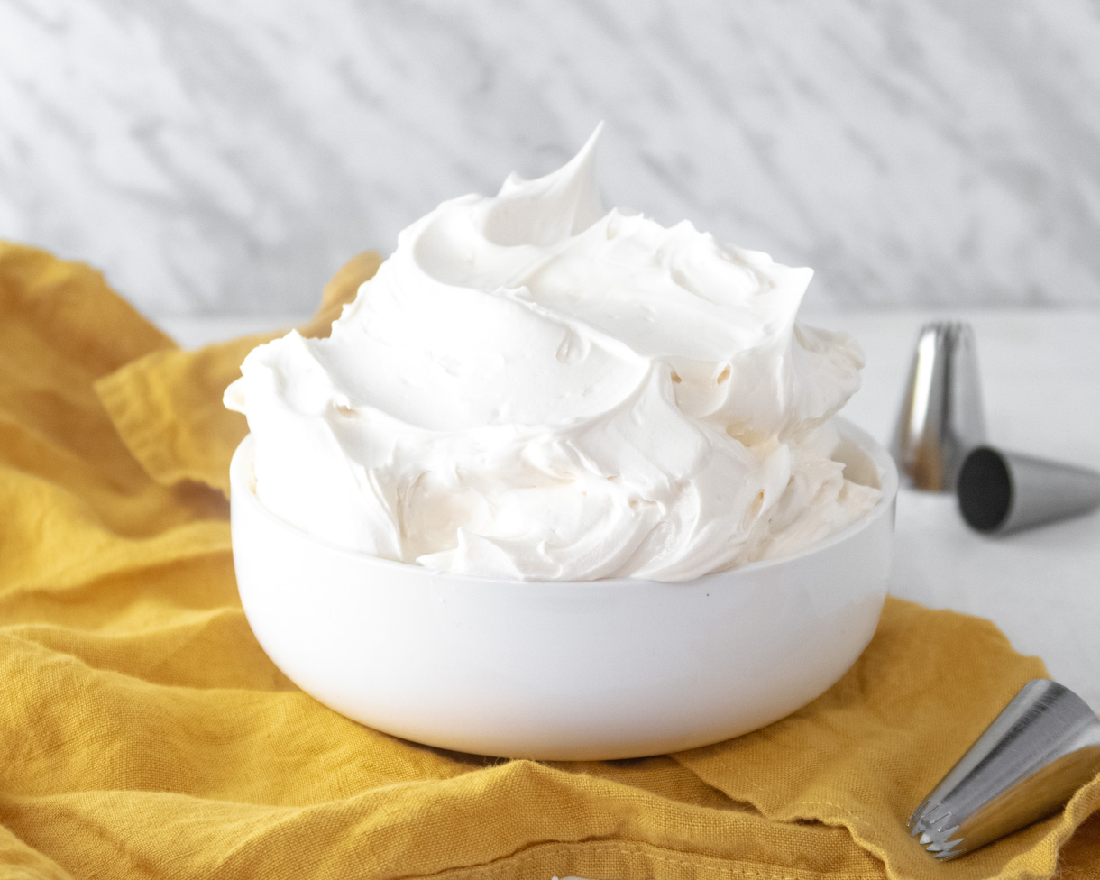 Vegan frosting in a white bowl on top of a mustard-coloured towel, with metal pastry tips around it.