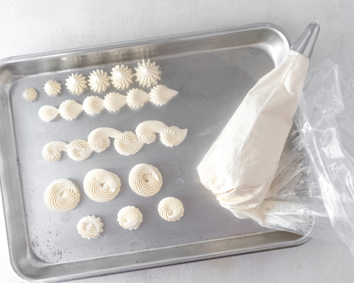 Vegan Italian meringue buttercream designs and borders piped on a silver baking tray with a filled pastry bag next to it.