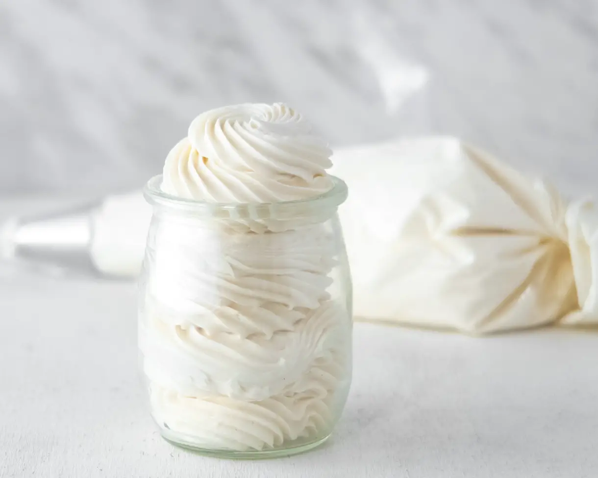 Vegan Italian meringue buttercream piped into a small glass jar with a pastry bag full of frosting in the background