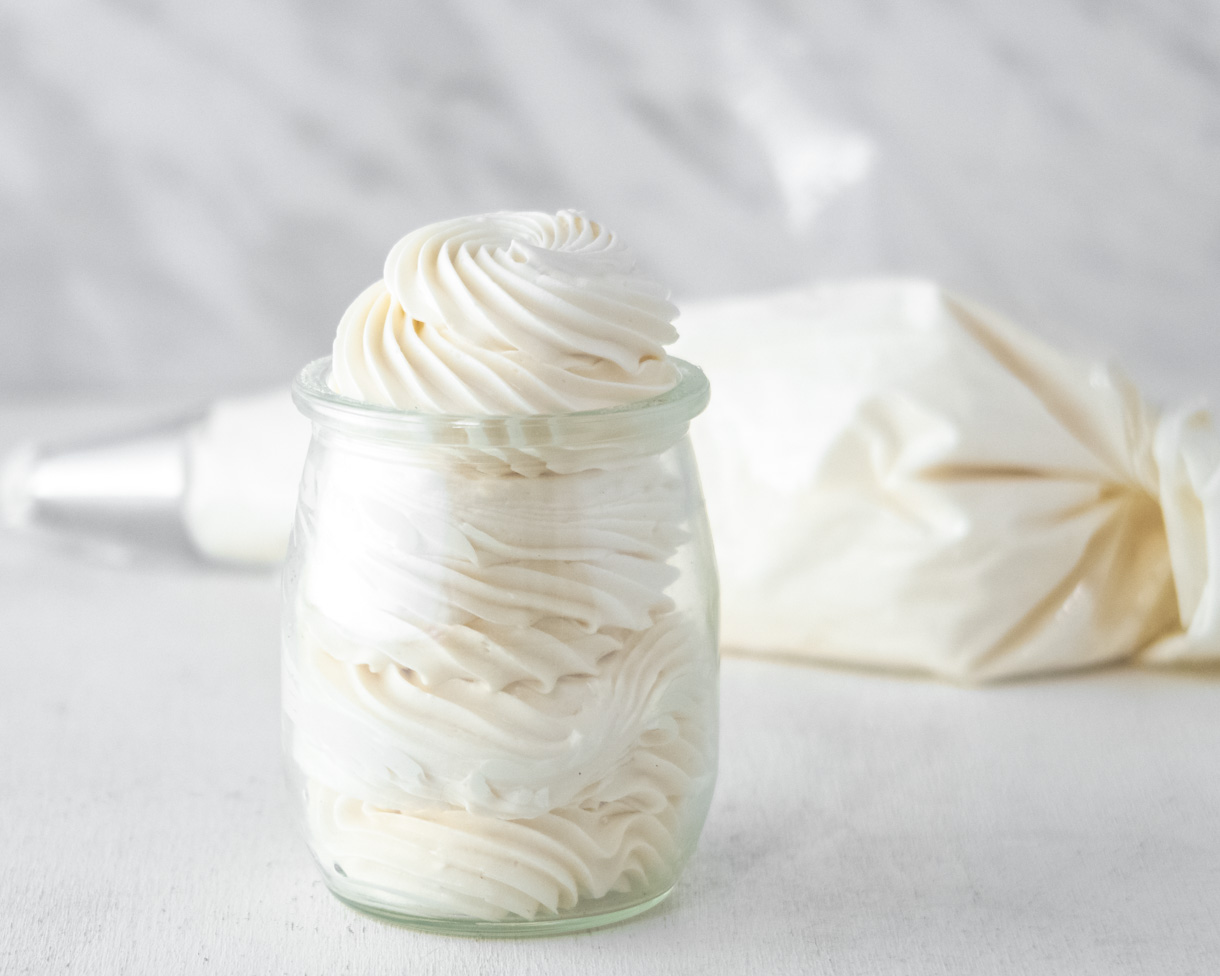 Vegan Italian meringue buttercream piped into a small glass jar with a pastry bag full of frosting in the background