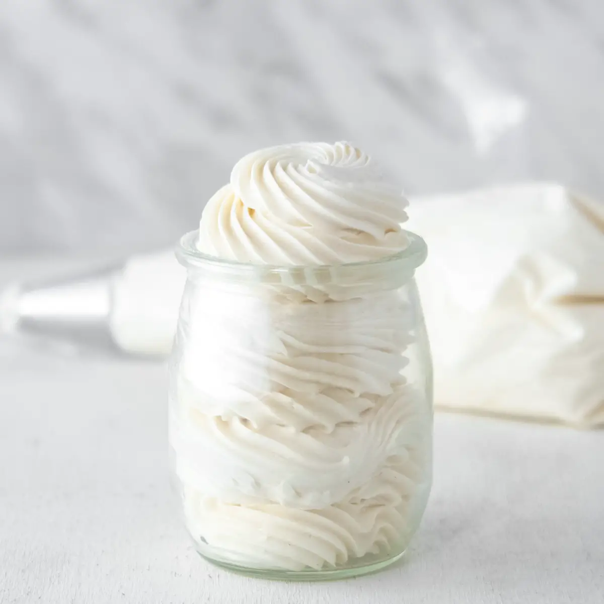Vegan Italian meringue buttercream piped into a small glass jar with a pastry bag full of frosting in the background