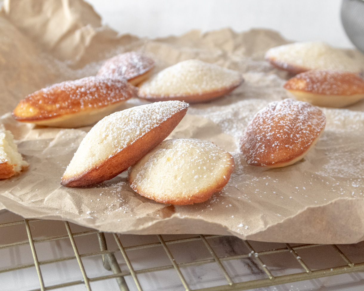 Madeleines lightly dusted with powdered sugar on a piece of kraft paper on a wire rack.