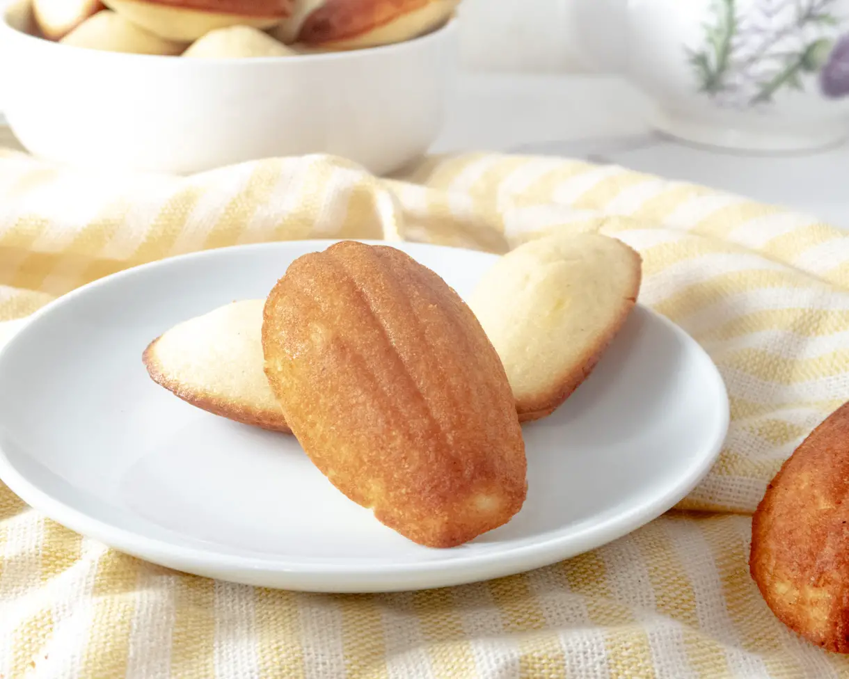 3 vegan madeleines on a small plate with a bowl of madeleines in the background.