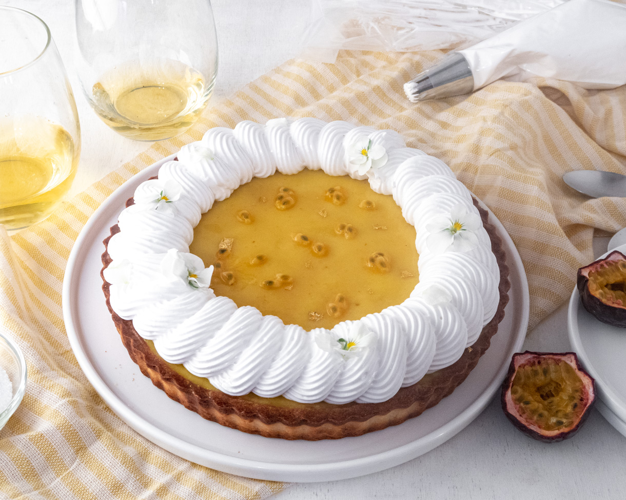 Vegan passionfruit meringue tart on a yellowed stripped tablecloth with wine glass and plates around it.