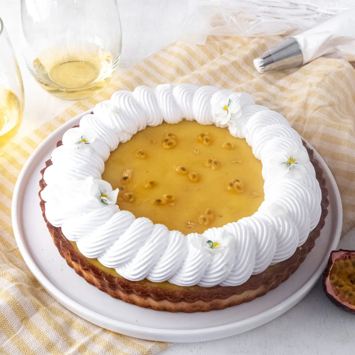 Vegan passionfruit meringue tart on a yellowed stripped tablecloth with wine glass and plates around it.
