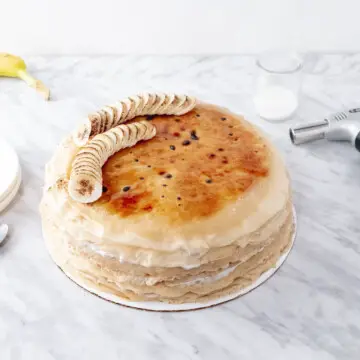 Vegan banana brulée mille crepes cake with a kitchen torch on the right and plates on the left.
