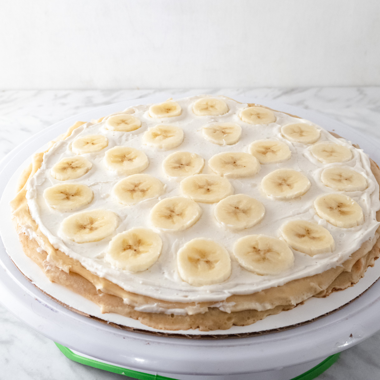 A crepe on a white cake board with a thin lawyer of diplomate cresm spread on it's surface and some sliced bananas placed all over the cream.