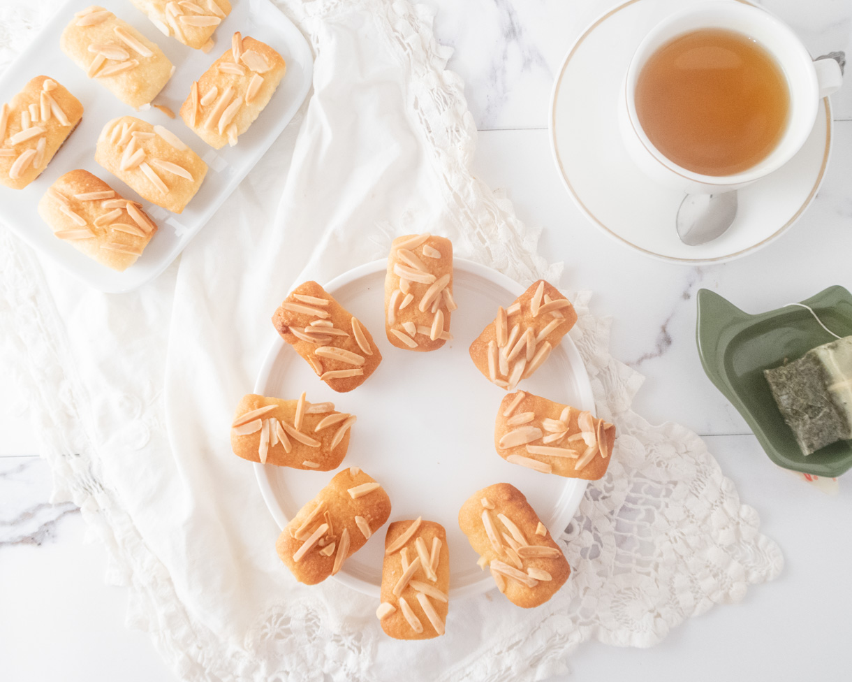 Plates filled with financiers on a white lace towel with a cup of tea and teabag on the top right corner