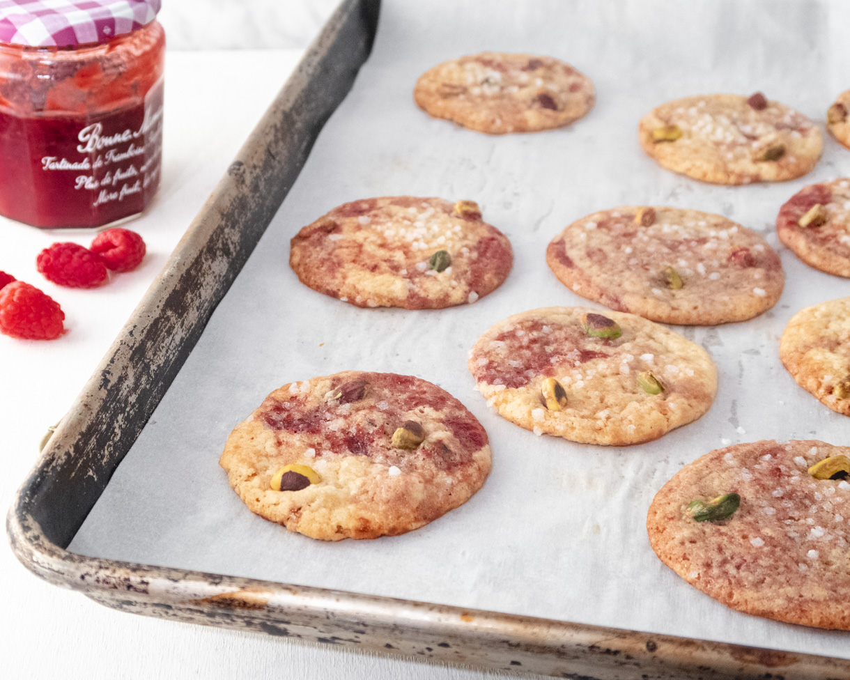 Cookies on a baking tray with jam and raspberry in the backgorund.