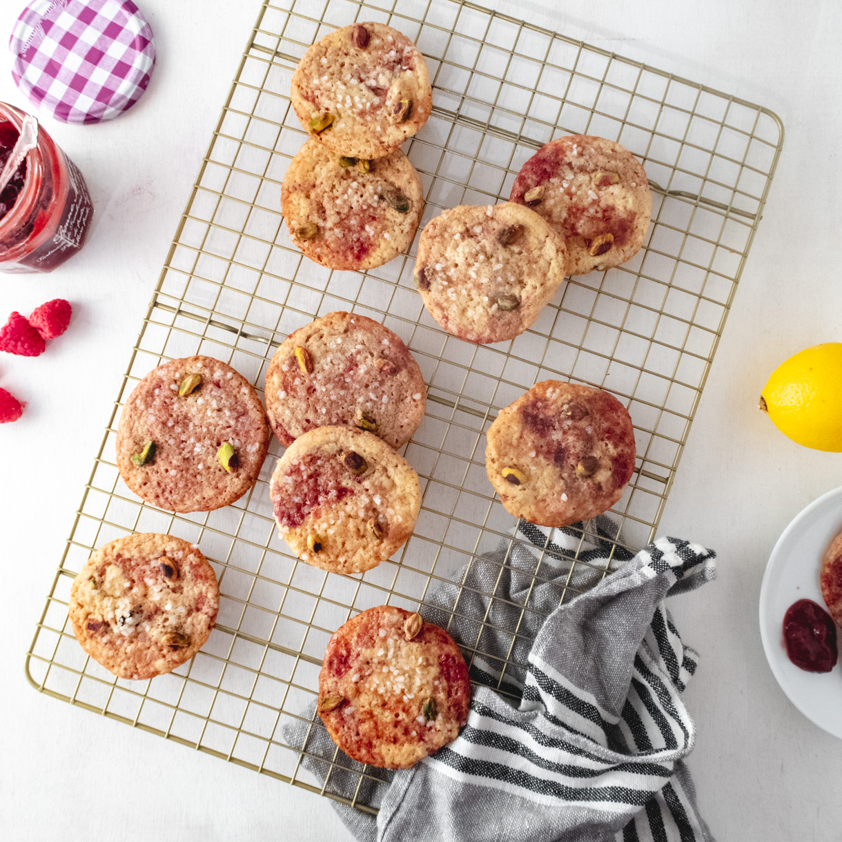 Vegan lemon pistachio cookies on a wire rack with jam, raspberries, a plate, and  a lemon around the rack.