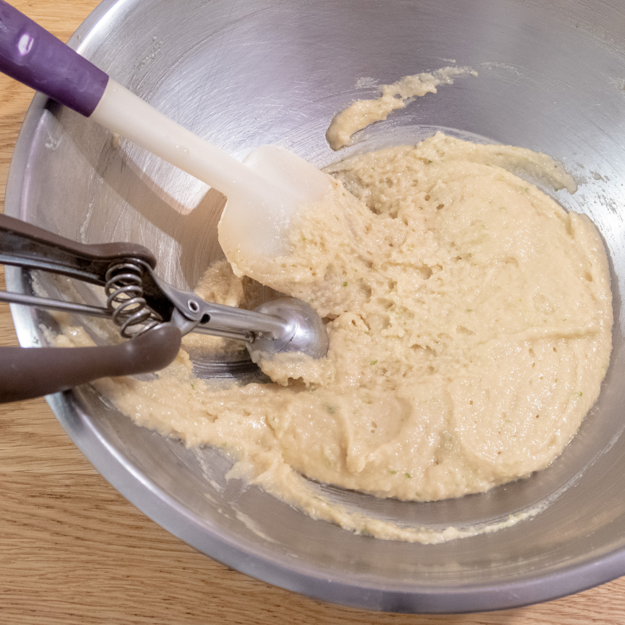 Finished financier batter in a large mixing bowl with a silicone spatule and small ice cream scoop in the bowl.