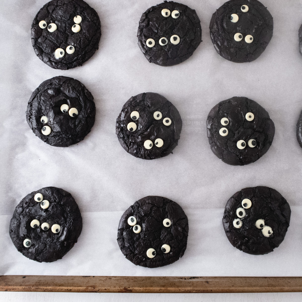 9 brownie cookies decorated with white chocolate eyes on a baking sheet lined with parchment paper.