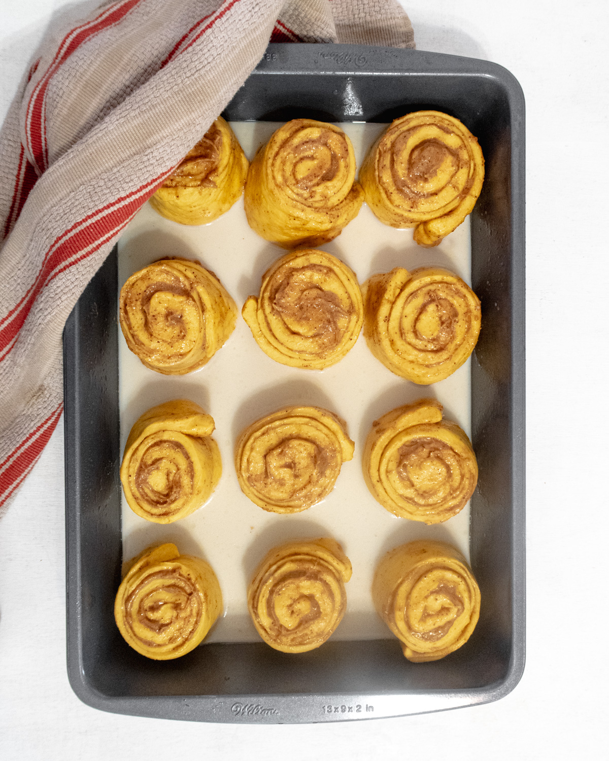 Raw unproofed cinnamon rolls in a baking pan with soy milk on the bottom.