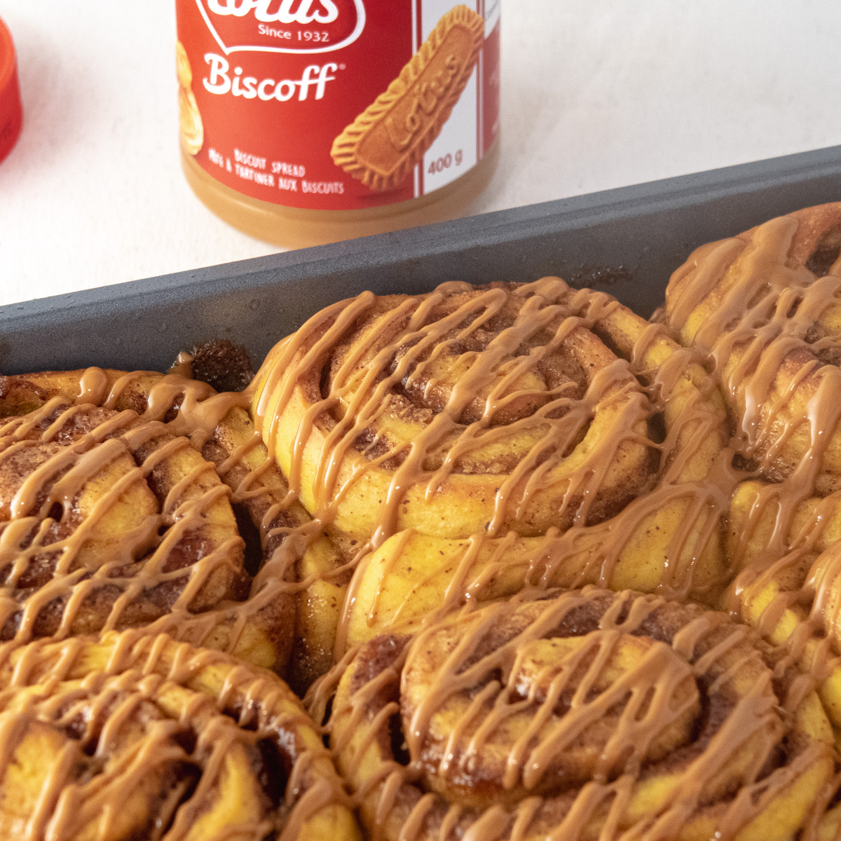 Close up of cookie butter drizzle on top of cinnamon buns with a jar of cookie butter in the background.