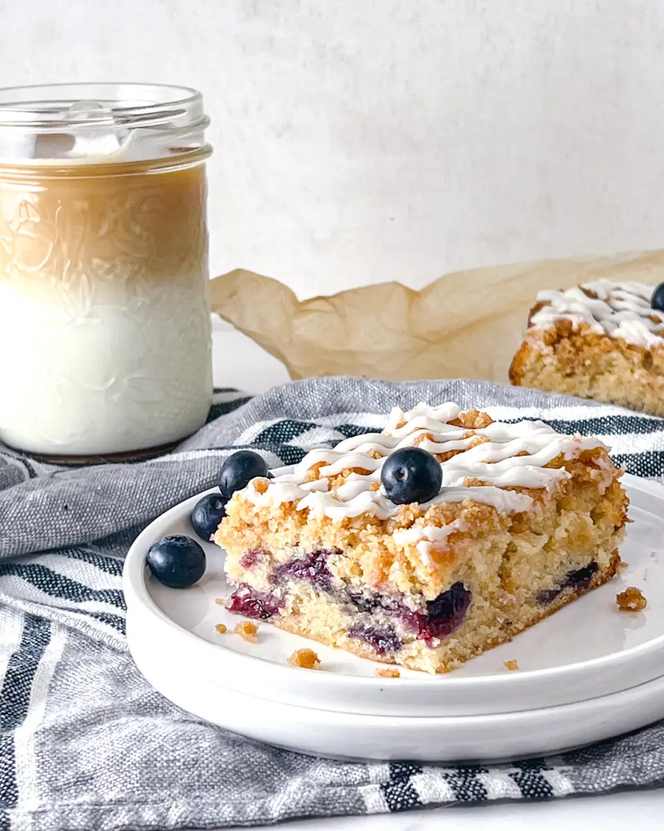 Side view of a piece of a piece of vegan lemon and blueberry coffee cake on a plate with an iced coffee in the background.